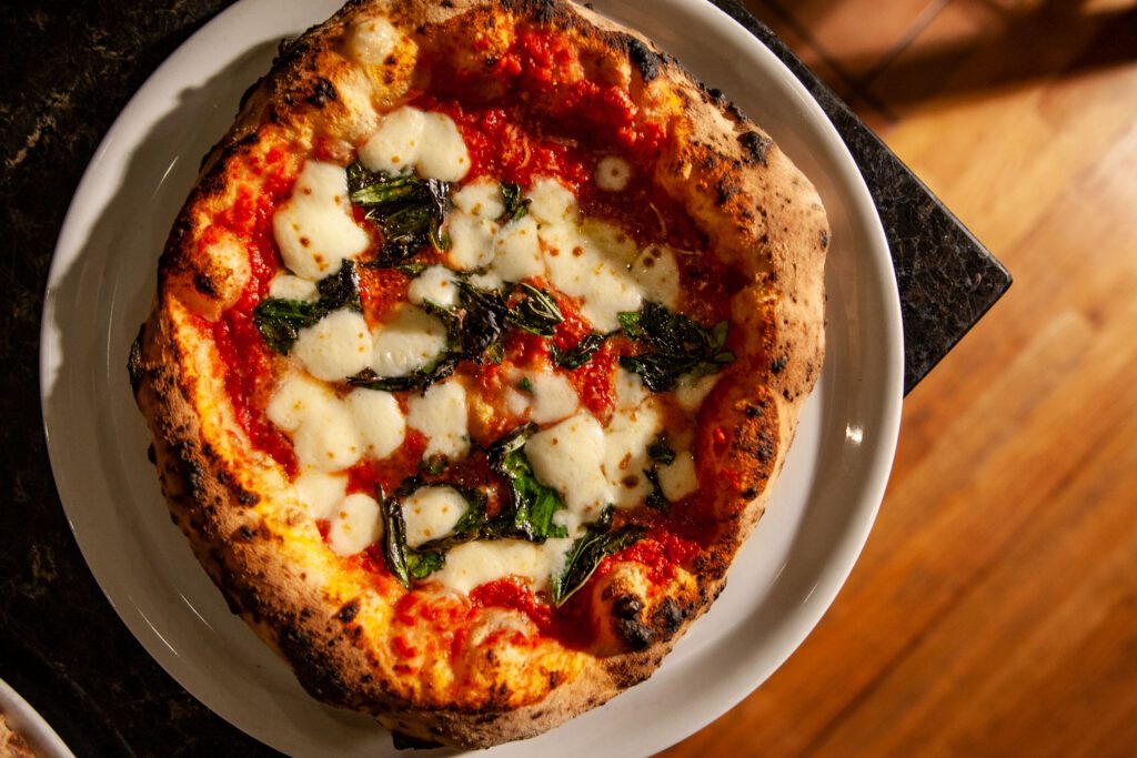 A Margherita pizza from Aquila Pizza Al Forno in Little Falls, NJ, featuring handmade mozzarella, San Marzano tomatoes, and fresh basil on a wood-fired crust with charred cornicione.