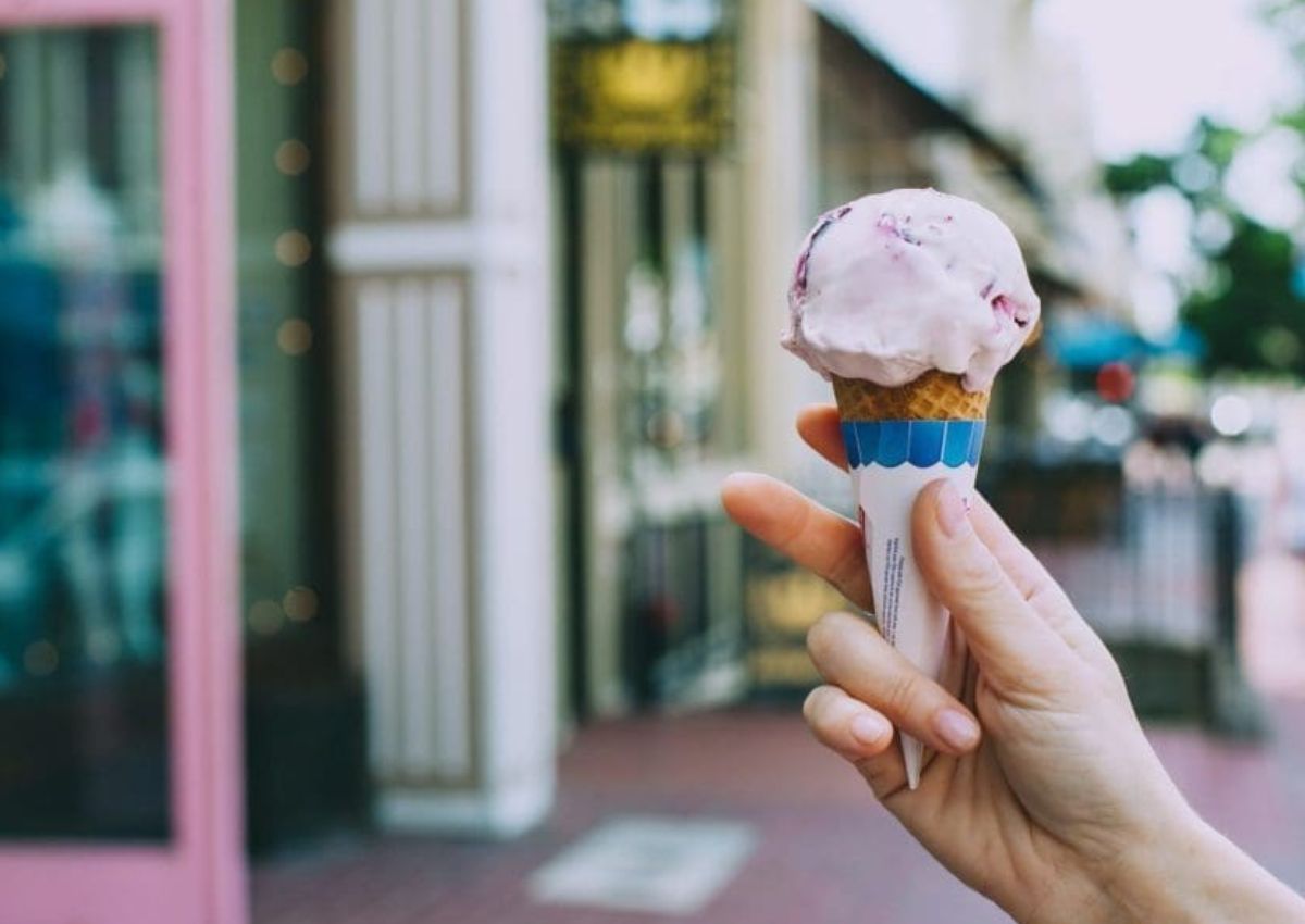 A scoop of ice cream on a waffle cone held outside an ice cream shop
