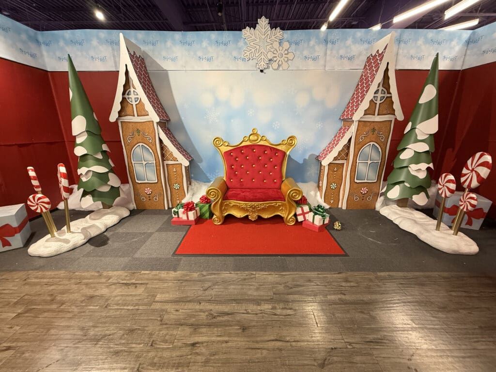 A gold-trimmed red throne sits among gingerbread houses, candy canes, snowy trees, and presents