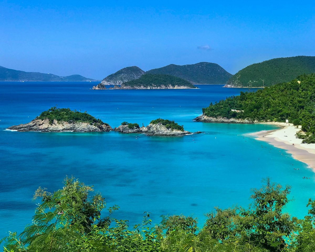 Aerial view of Trunk Bay in St. John, U.S. Virgin Islands, showing turquoise Caribbean water, white sand beach, and small rocky islands surrounded by lush green hills