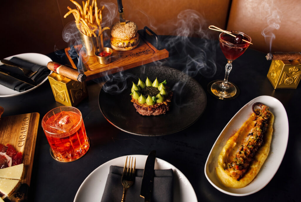 a spread of food on the spring menu with a cocktail and lit cigar at Cigarro in Hackensack, NJ