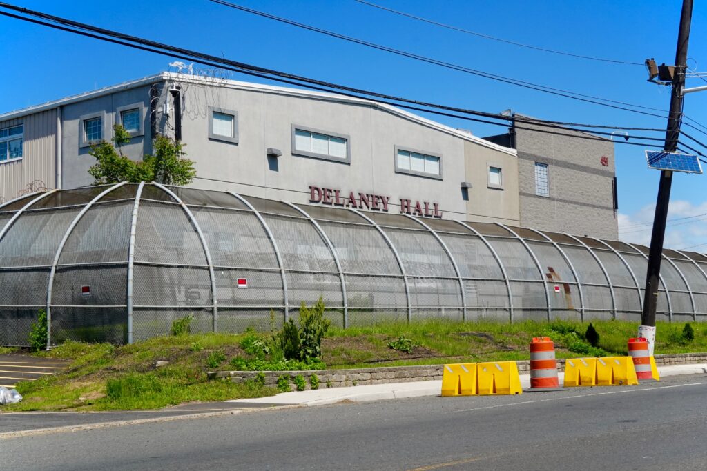 The exterior of Delaney Hall ICE detention facility in Newark, New Jersey, with security fencing and road barriers in the foreground