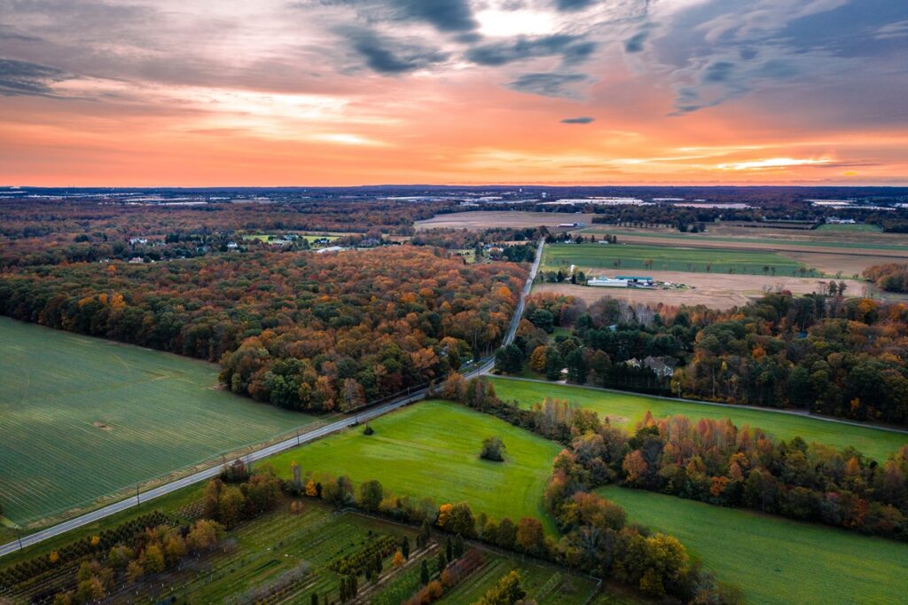 Aerial view of New Jersey farmland and orchards at sunset, with fields bordering suburban development and warehouses on the horizon.